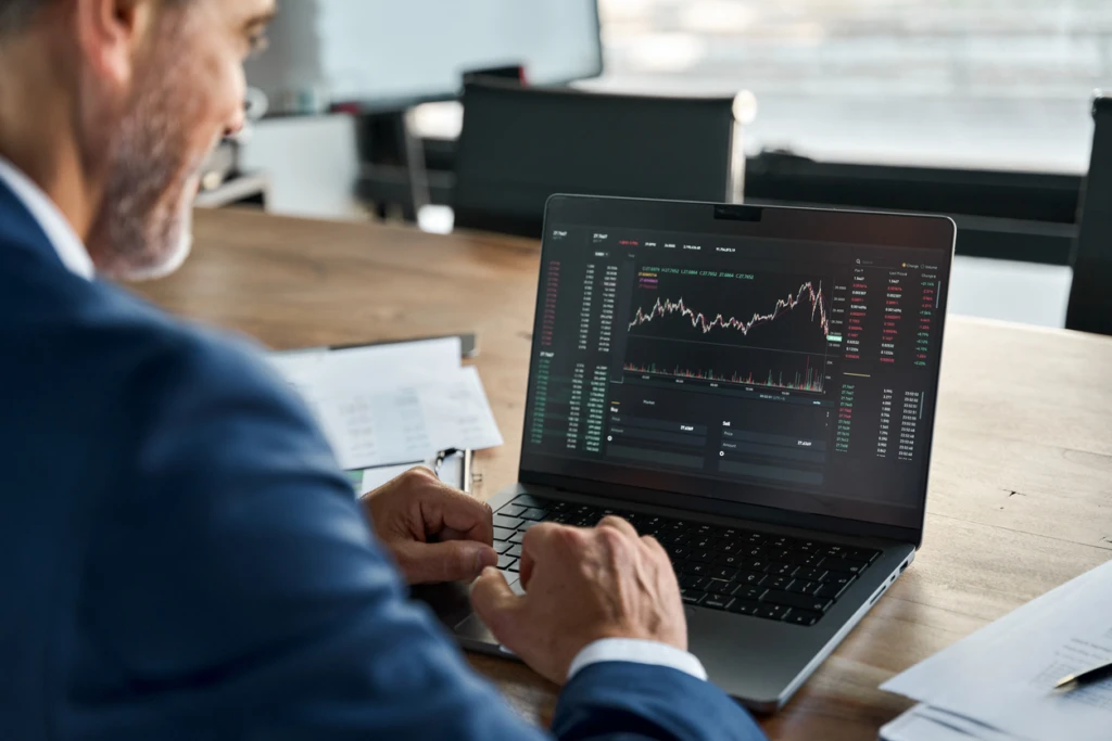 Homem analisando gráficos financeiros no computador em um escritório moderno, com foco em investimentos e mercado financeiro.