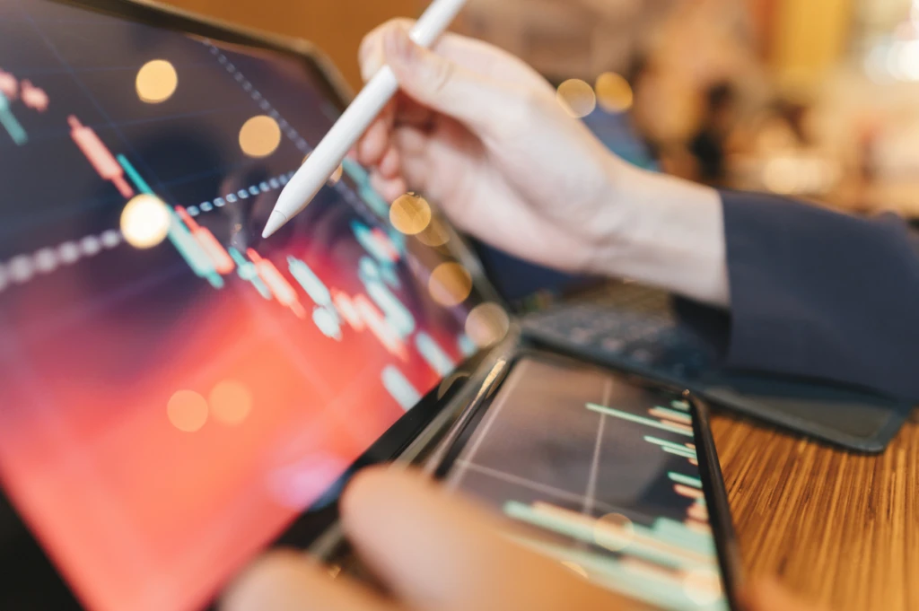 Pessoa analisando gráficos de investimentos em dispositivos móveis com uso de caneta em ambiente financeiro.