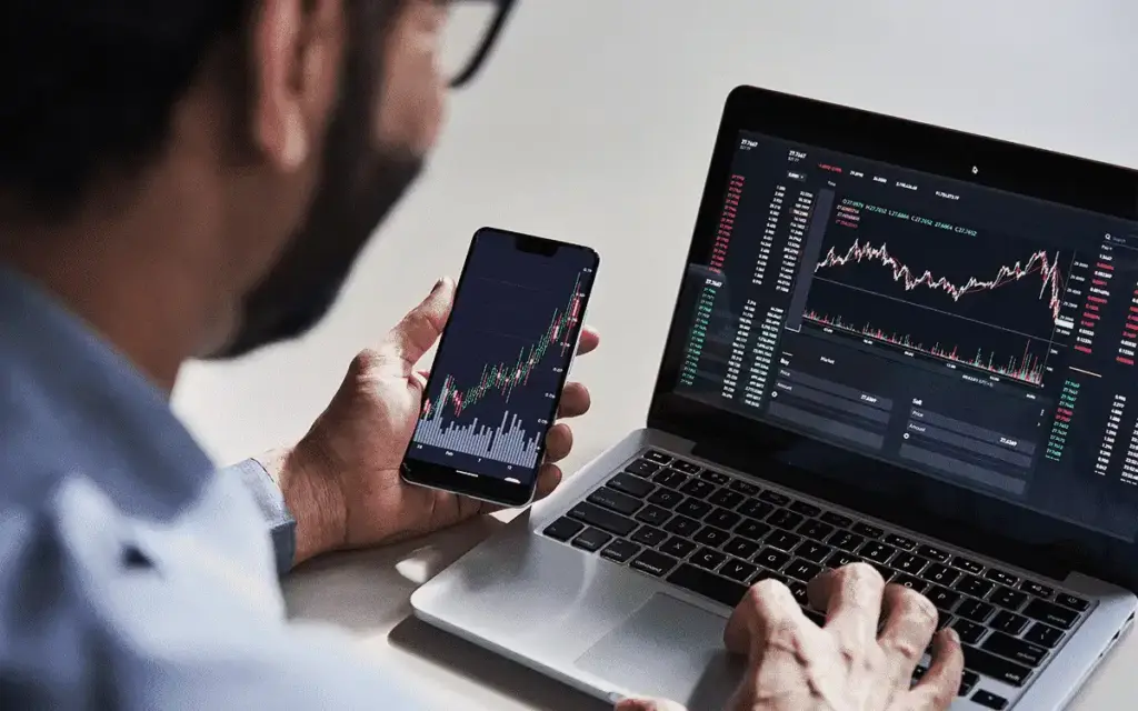 Homem analisando gráficos de ações em smartphone e notebook, monitorando mercado financeiro e tendências de investimentos.