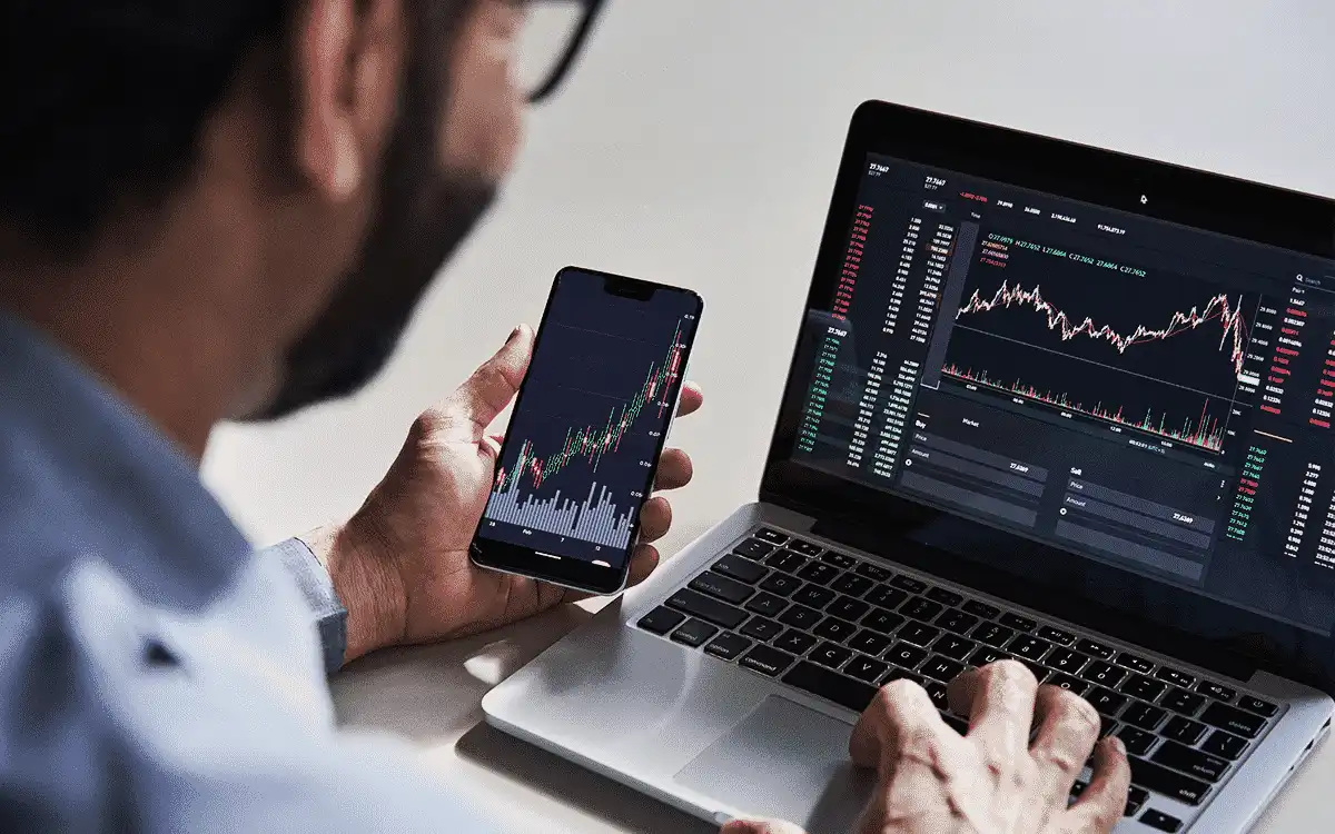 Homem analisando gráficos de ações em smartphone e notebook, monitorando mercado financeiro e tendências de investimentos.