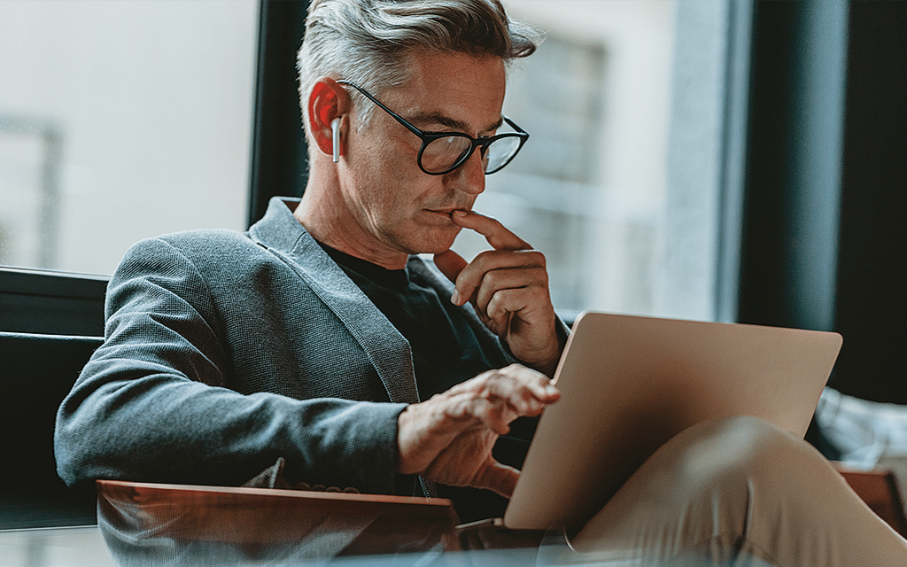Homem de terno e óculos usando um tablet, pensativo, sentado perto de uma janela em um ambiente de escritório moderno, promovendo produtividade e tecnologia.