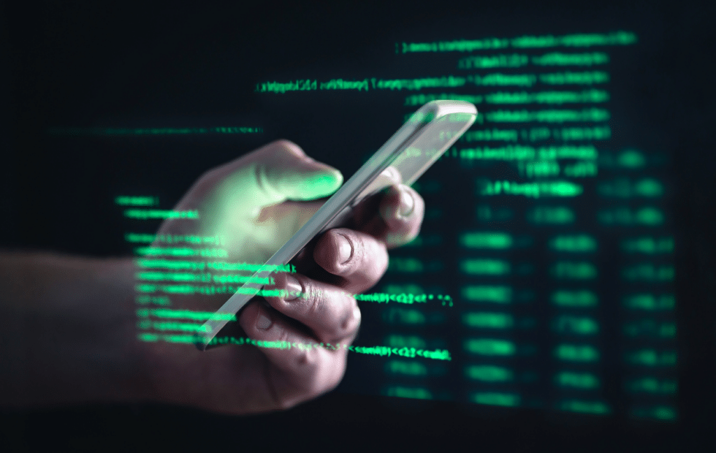 Imagem de uma mão segurando um smartphone com códigos de programação verdes ao redor, simbolizando tecnologia, digital e segurança cibernética.