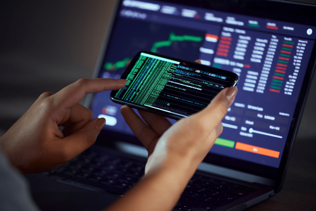Pessoa analisando gráficos financeiros e dados de bolsa de valores no smartphone e computador, rotina de investimentos e mercado financeiro.