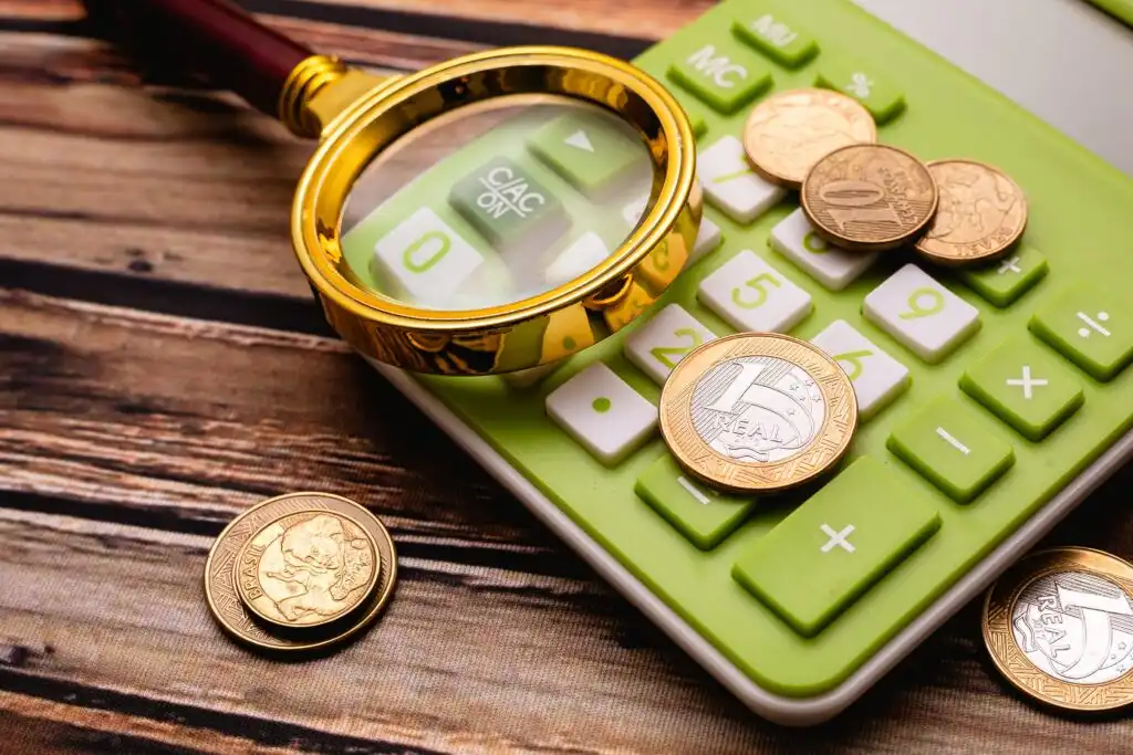 Imagem de uma calculadora verde com moedas de euro e uma lupa de ouro sobre ela, sobre uma mesa de madeira, representando finanças e economia.