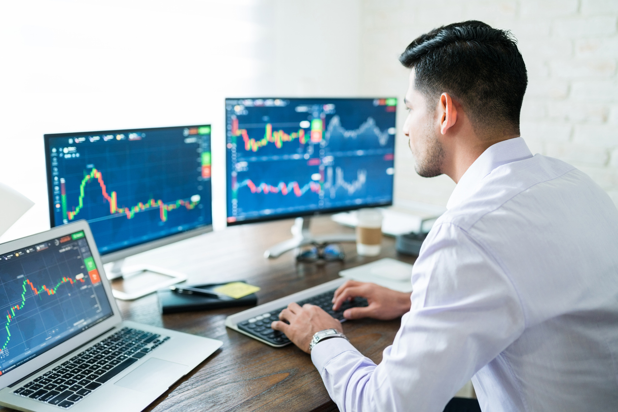 Homem trabalhando com múltiplos monitores exibindo gráficos e análises de ações, focado em investimento e mercado financeiro.
