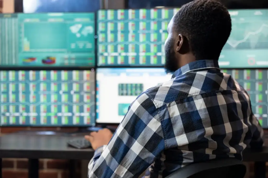 Homem trabalhando em frente a múltiplos monitores exibindo gráficos e dados financeiros, análise de mercado e tecnologia financeira.