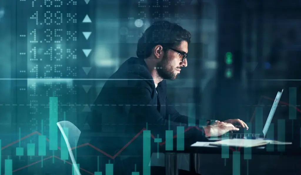 Homem de terno e óculos trabalhando em um laptop com gráficos de análise financeira e mercado de ações ao fundo, representando investimentos e análise de mercado.