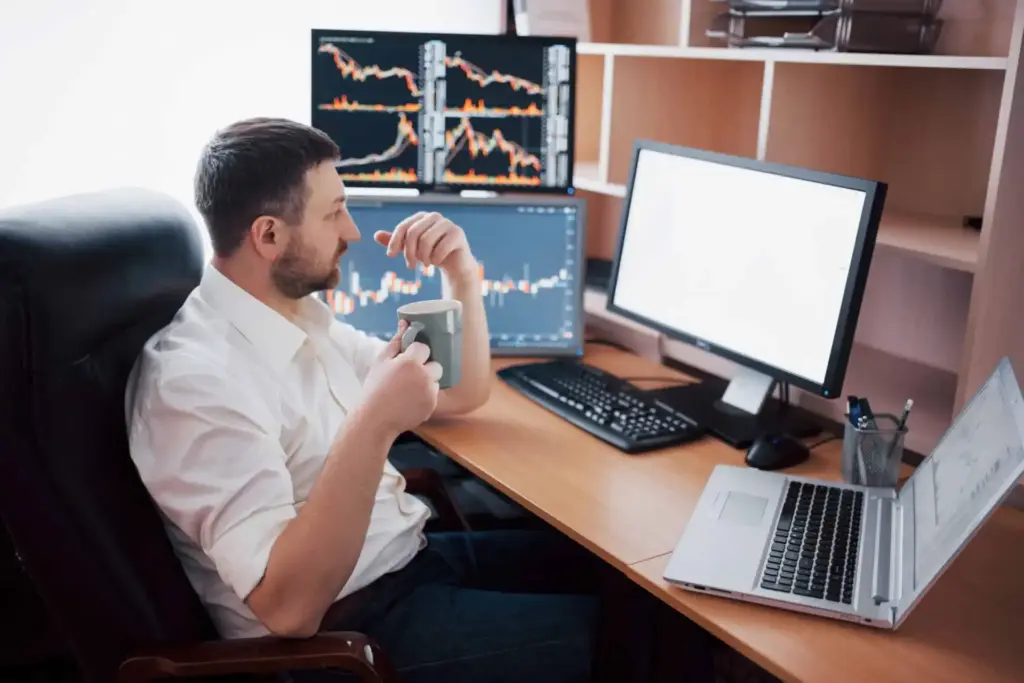 Homem analisando gráficos financeiros em várias telas de computador em um escritório, com foco em mercado de ações.