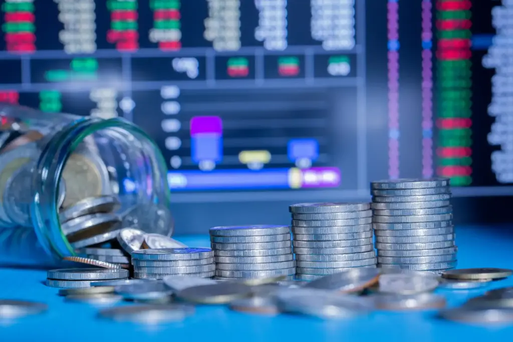 Investimento em ações e mercado financeiro: pilhas de moedas empilhadas e vidro com moedas, fundos de tela com gráficos de bolsa de valores ao fundo, simbolizando crescimento financeiro e investimento.