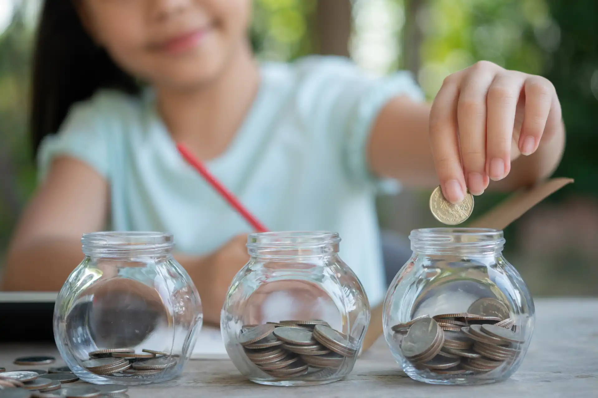 Criança colocando moeda em pote de vidro, incentivando economia infantil ao ar livre.