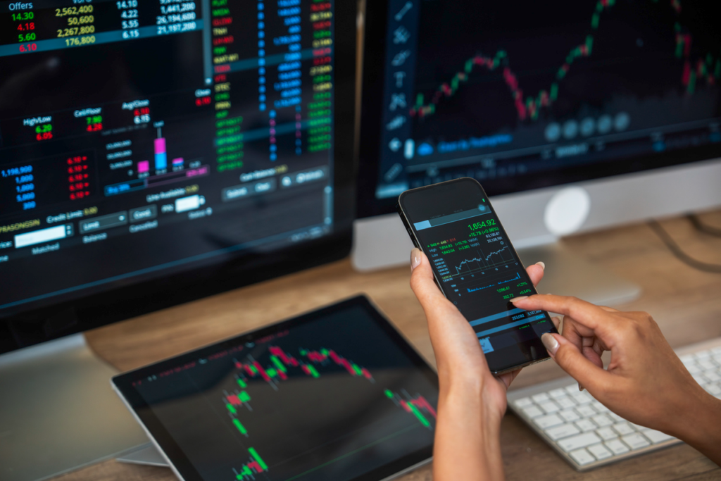 Pessoa analisando gráficos de mercado financeiro em vários dispositivos eletrônicos, incluindo computador, tablet e smartphone, com foco em dados e tendências de ações e investimentos.