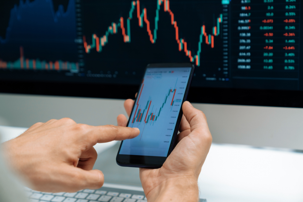 Pessoa analisando gráficos financeiros em um smartphone, com tela de computador ao fundo exibindo dados de mercado e investimentos, símbolo de análise de ações.