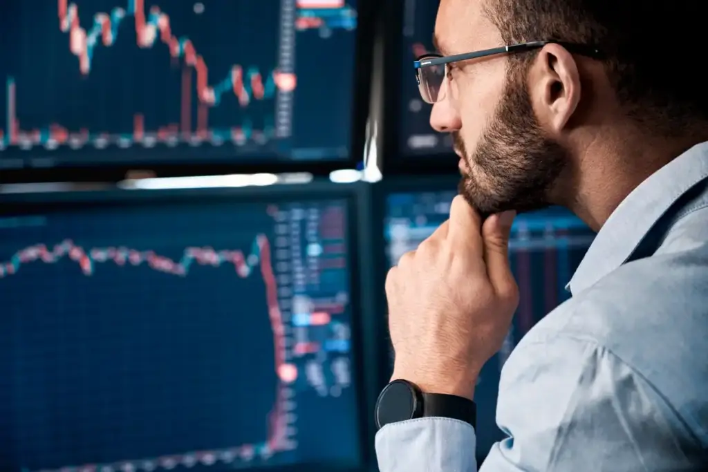 Homem analisando múltiplos gráficos de ações na tela de computador, indicando estudo de mercado financeiro e investimentos