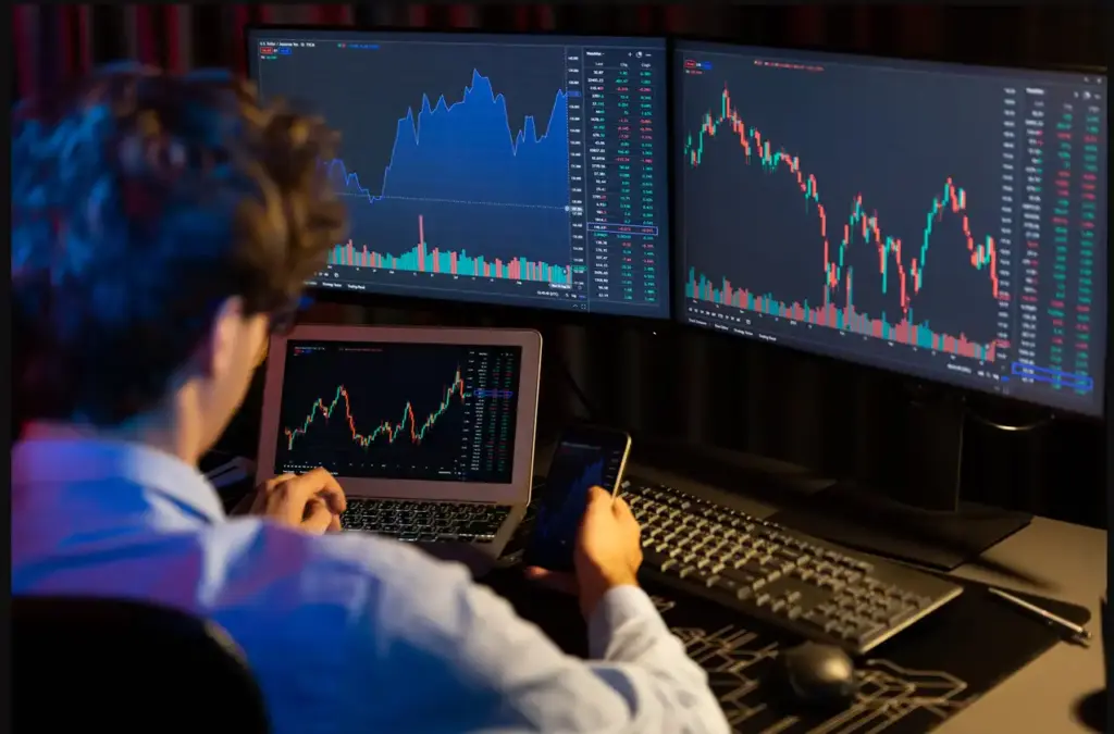 Pessoa analisando gráficos de mercado financeiro em múltiplos monitores e smartphone, com foco em investimentos e trading.