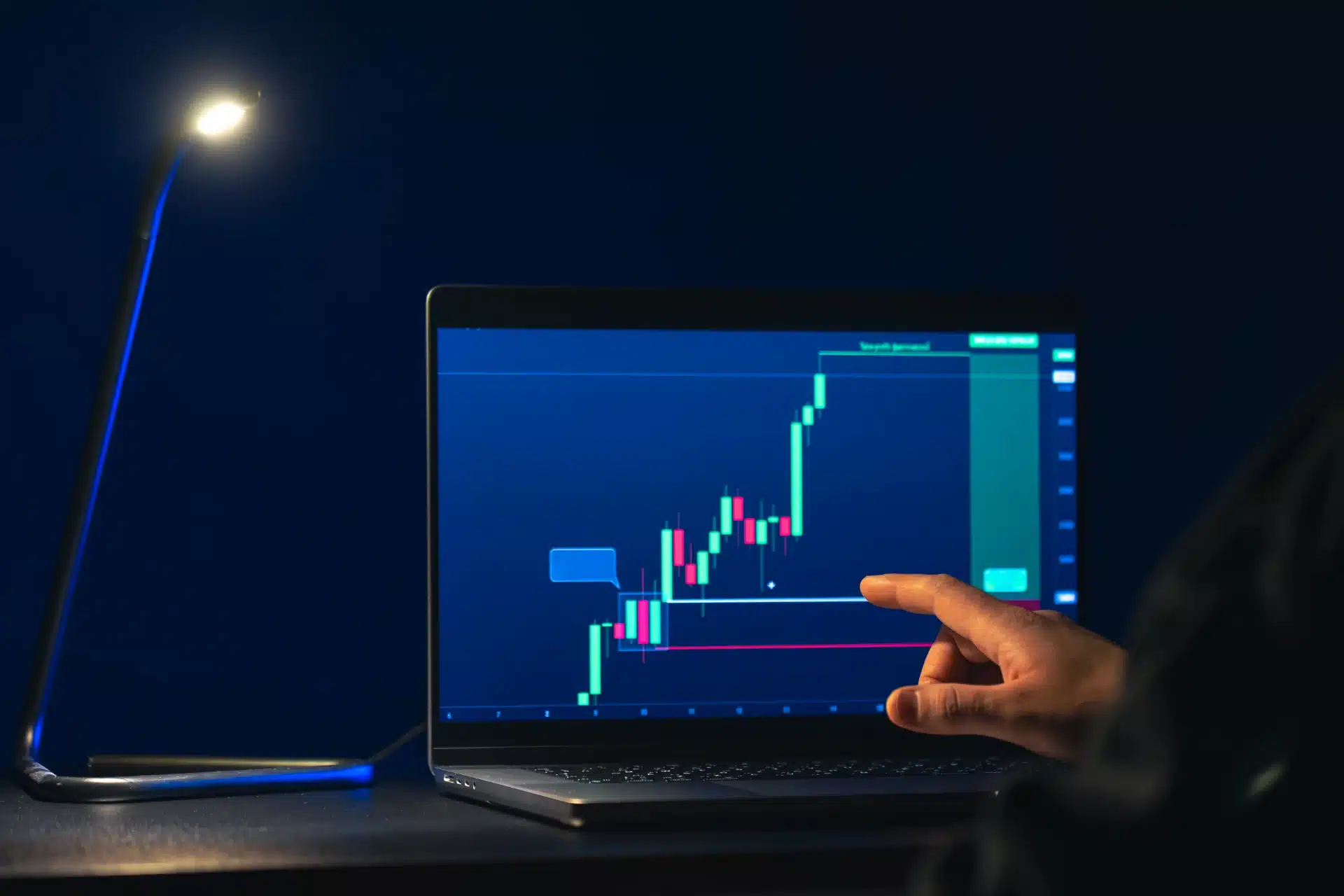 Pessoa analisando gráficos de investimentos em um laptop com uma luminária ao lado, símbolos de crescimento financeiro e trading.