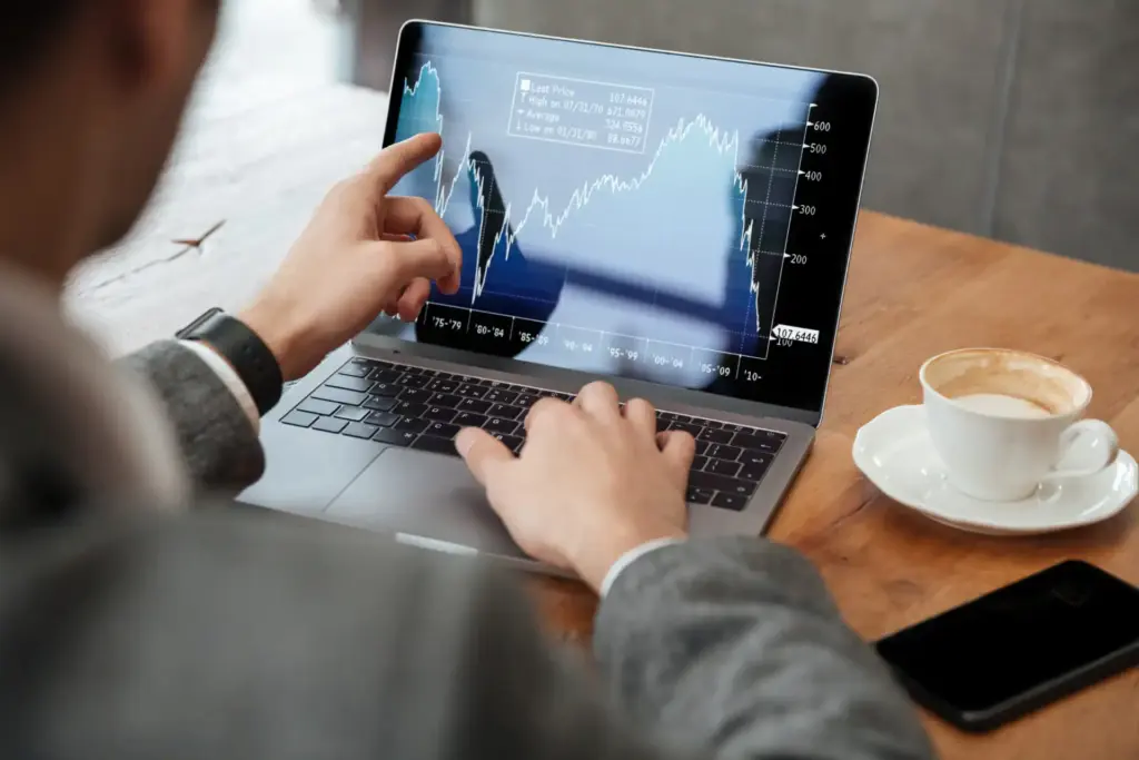 Pessoa analisando gráficos de mercado de ações em um computador portátil em uma cafeteria, destacando movimentos de alta e baixa na bolsa.