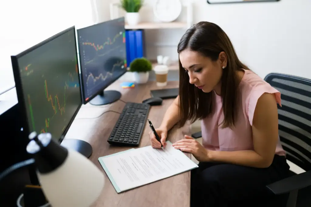 Mulher analista financeira trabalhando em escritório com dois monitores exibindo gráficos de investimentos, papel, caneta e computador.