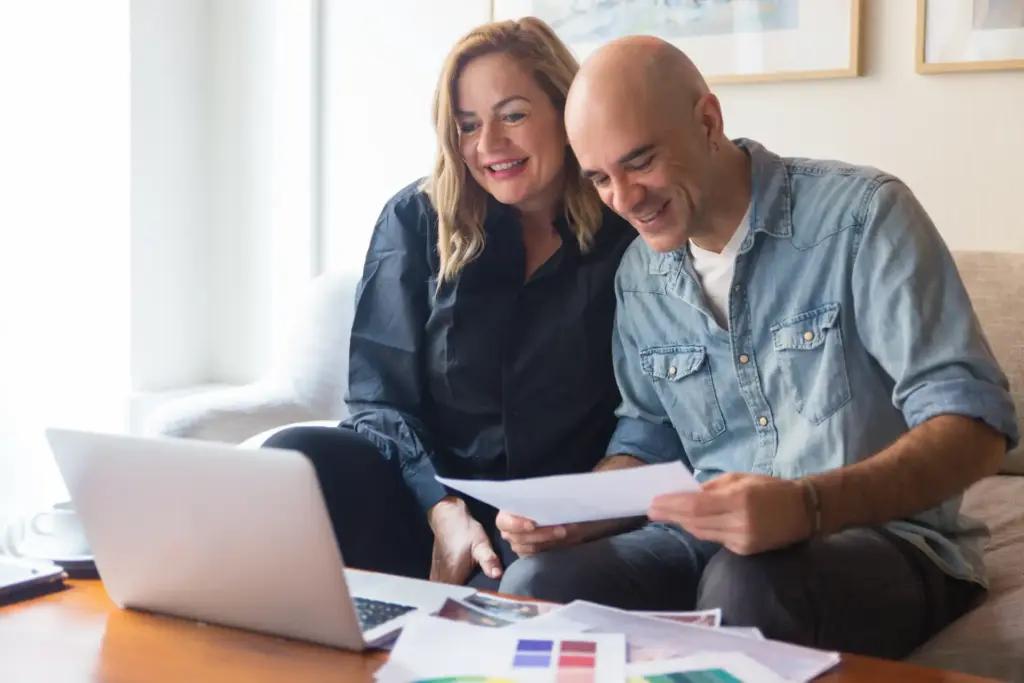 Casal feliz analisando documentos e usando laptop na sala de estar, expressão de satisfação, ambiente acolhedor e iluminado.