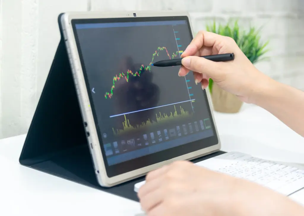 Pessoa analisando gráficos de mercado financeiro em um tablet com caneta, com foco em investimentos e ações, ambiente de escritório.