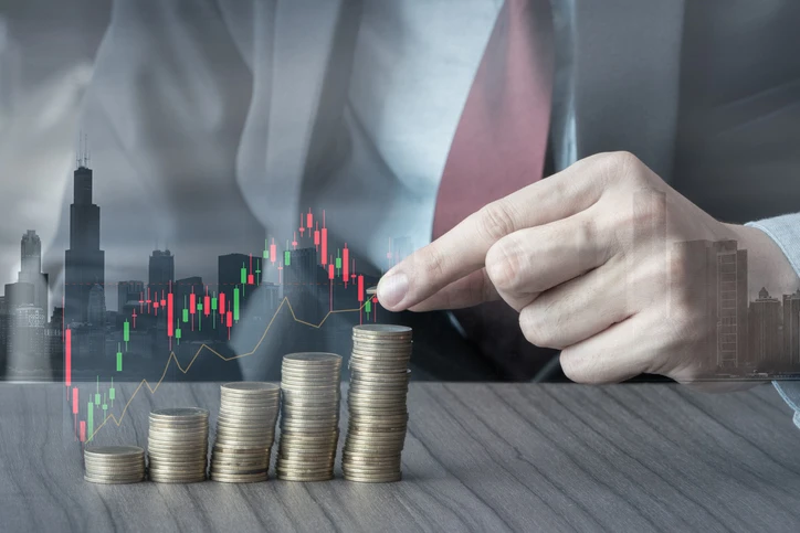 Homem de terno fazendo uma análise financeira com gráficos de mercado e pilhas de moedas, simbolizando crescimento econômico e investimentos.