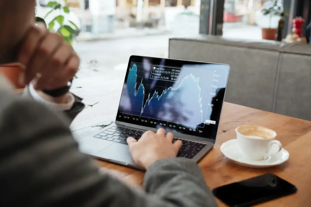 Pessoa analisando gráficos financeiros em um laptop em uma cafeteria, com uma xícara de café ao lado, durante o dia.