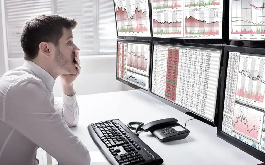 Homem analisando gráficos de ações em múltiplos monitores de computador, demonstrando atenção aos detalhes no mercado financeiro.