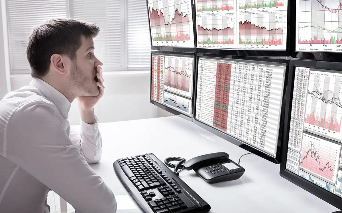Homem analisando gráficos de ações em múltiplos monitores de computador, demonstrando atenção aos detalhes no mercado financeiro.