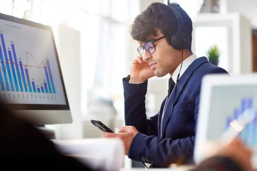 Homem de negócios usando headset, analisando gráficos e dados financeiros em ambiente de escritório, simbolizando análise financeira e investimentos.