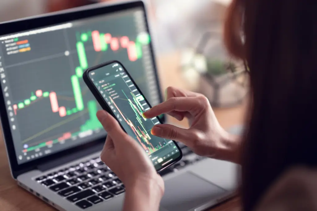 Pessoa usando smartphone para analisar gráficos de investimentos financeiros com tela de computador ao fundo