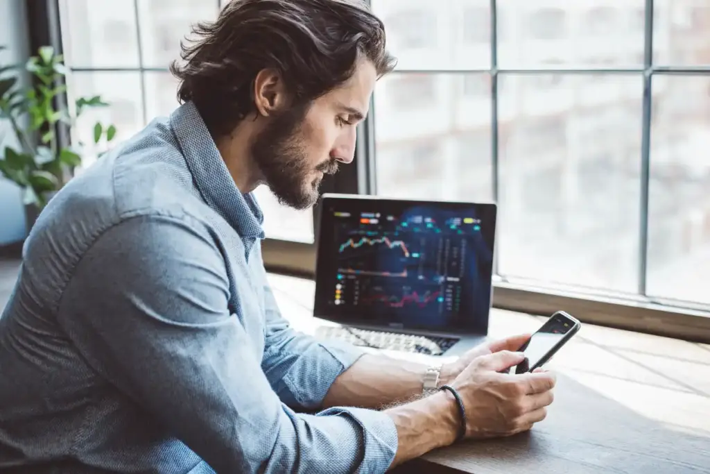 Homem analisando gráficos financeiros em um escritório moderno enquanto usa um smartphone, com computador exibindo dados de mercado ao fundo.