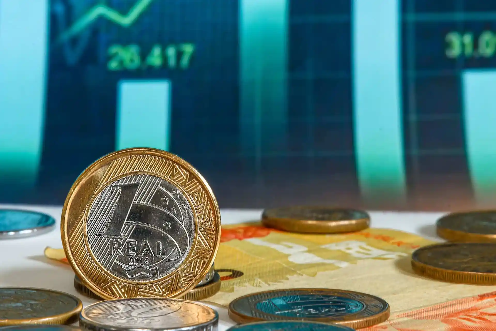 Imagem de uma moeda de 1 real brasileira e outras moedas de diferentes valores em destaque, com gráficos de crescimento financeiro ao fundo, representando investimentos e economia.