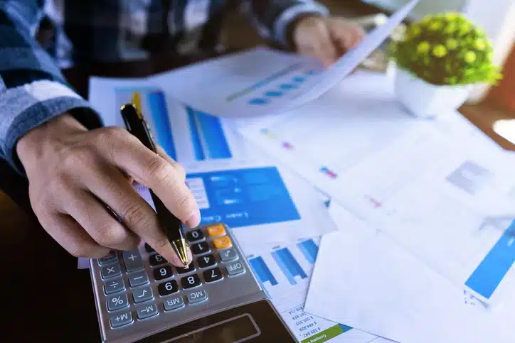 Pessoa analisando documentos e utilizando uma calculadora em um ambiente de trabalho para fins financeiros ou de negócios, com gráficos coloridos ao fundo.