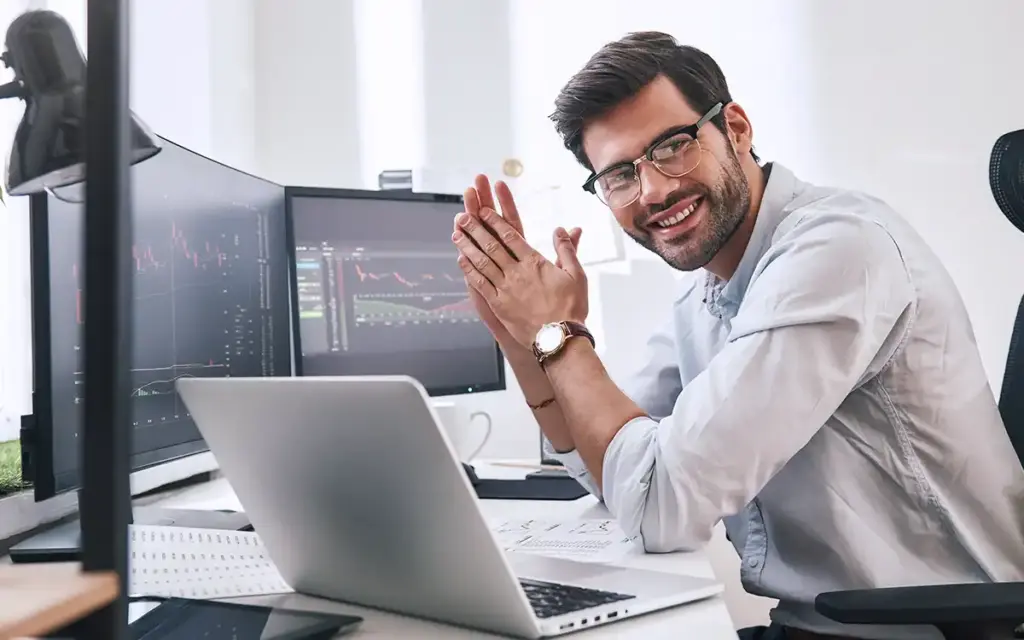 Homem sorridente trabalhando com múltiplos monitores de computador exibindo gráficos financeiros, indicando sucesso em investimentos ou análise de mercado.