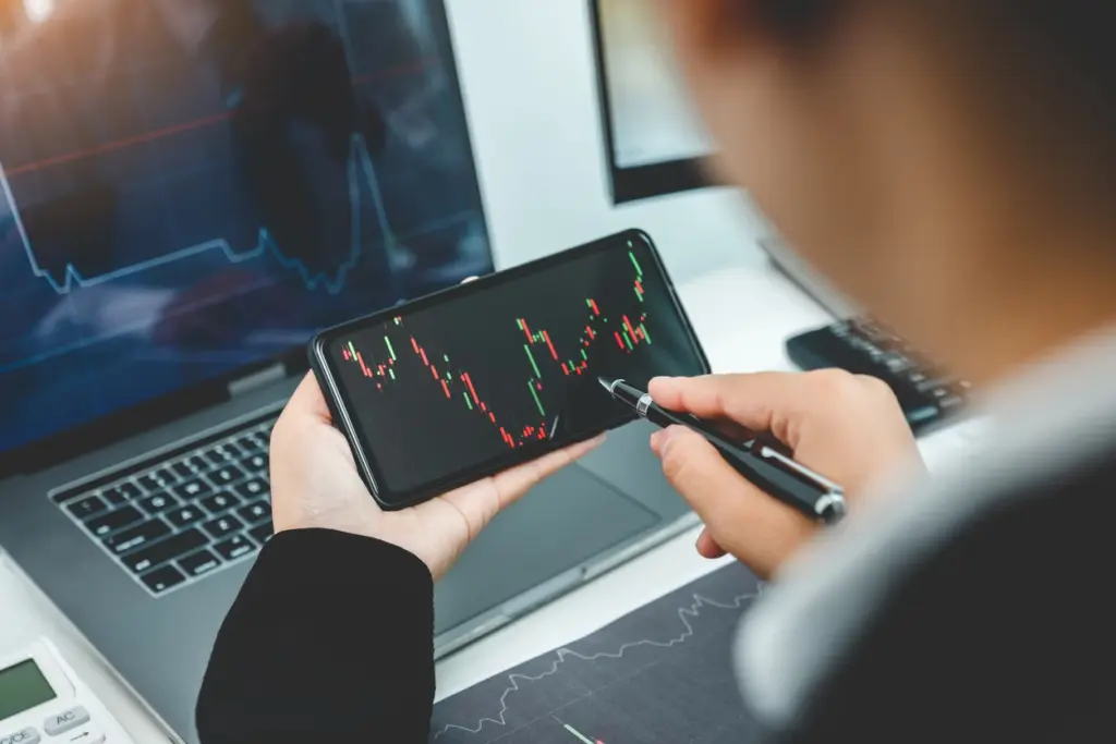 Pessoa analisando gráficos de ações no smartphone, computador e monitor, representando investimento em Bolsa de Valores.