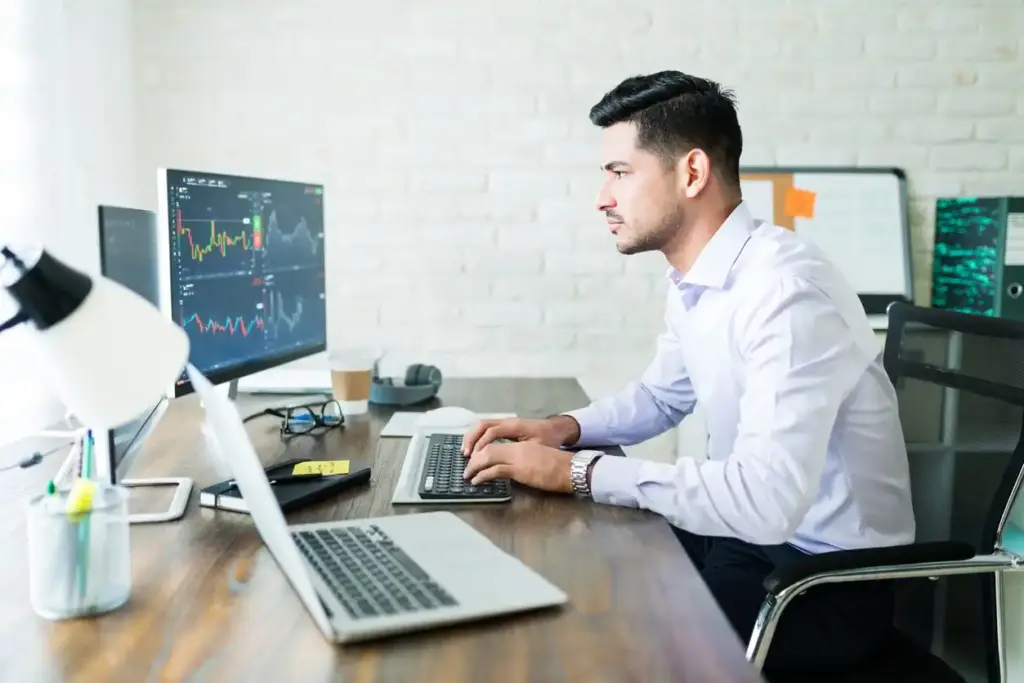 Homem trabalhador analisando gráficos de mercado financeiro em seu computador no escritório moderno, com várias telas exibindo dados de investimentos.