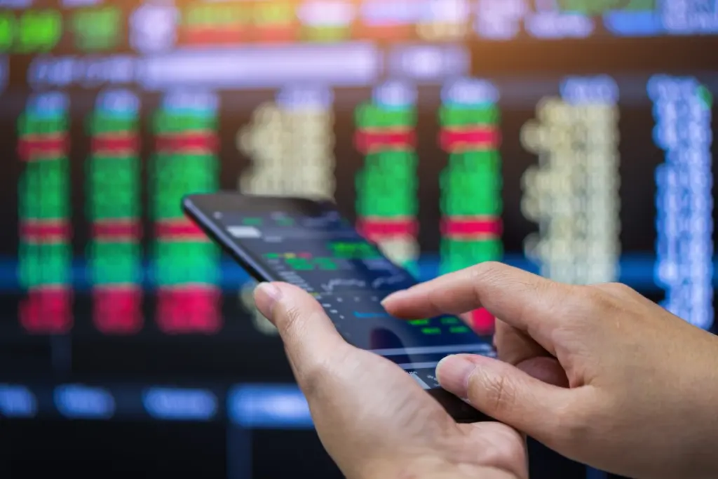 Pessoa usando smartphone para acompanhar dados de mercado financeiro com tela de ações de fundo em uma tela de computador.
