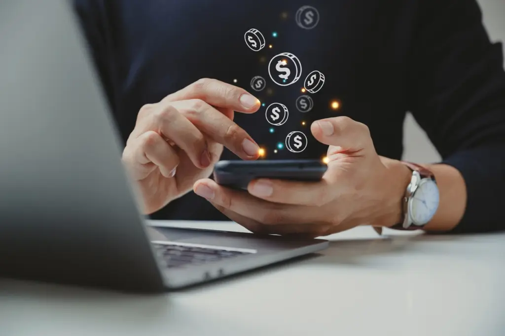 Pessoa usando smartphone enquanto moedas e símbolos de dinheiro flutuam na tela, sugerindo pagamento digital, transações e gestão financeira em ambiente de escritório.
