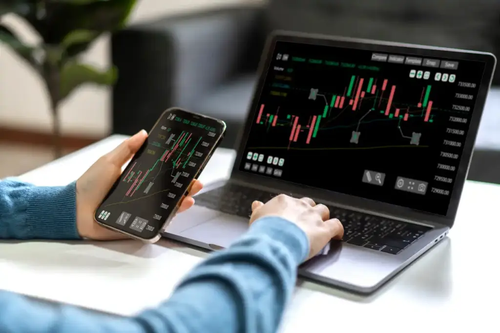 Pessoa analisando gráficos de ações em smartphone e computador, com foco em investimentos e mercado financeiro.