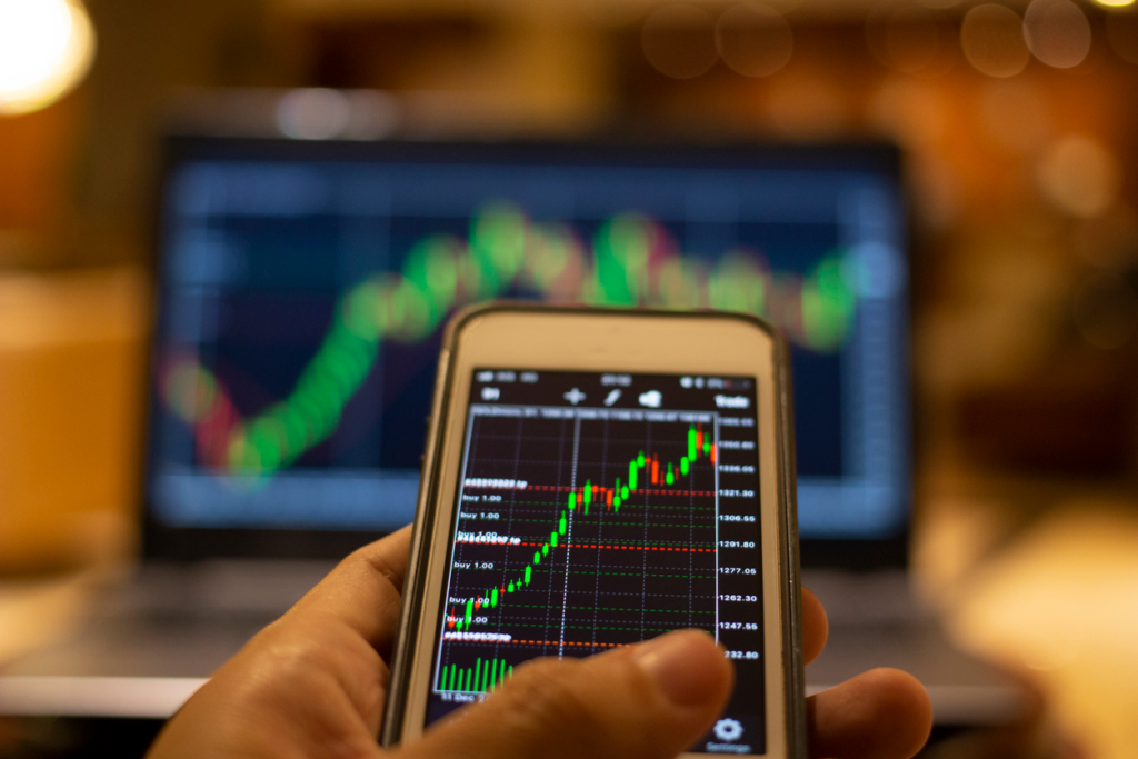 Pessoa analisando gráficos de mercado de ações em smartphone e computador portátil, representando investimentos e finanças.