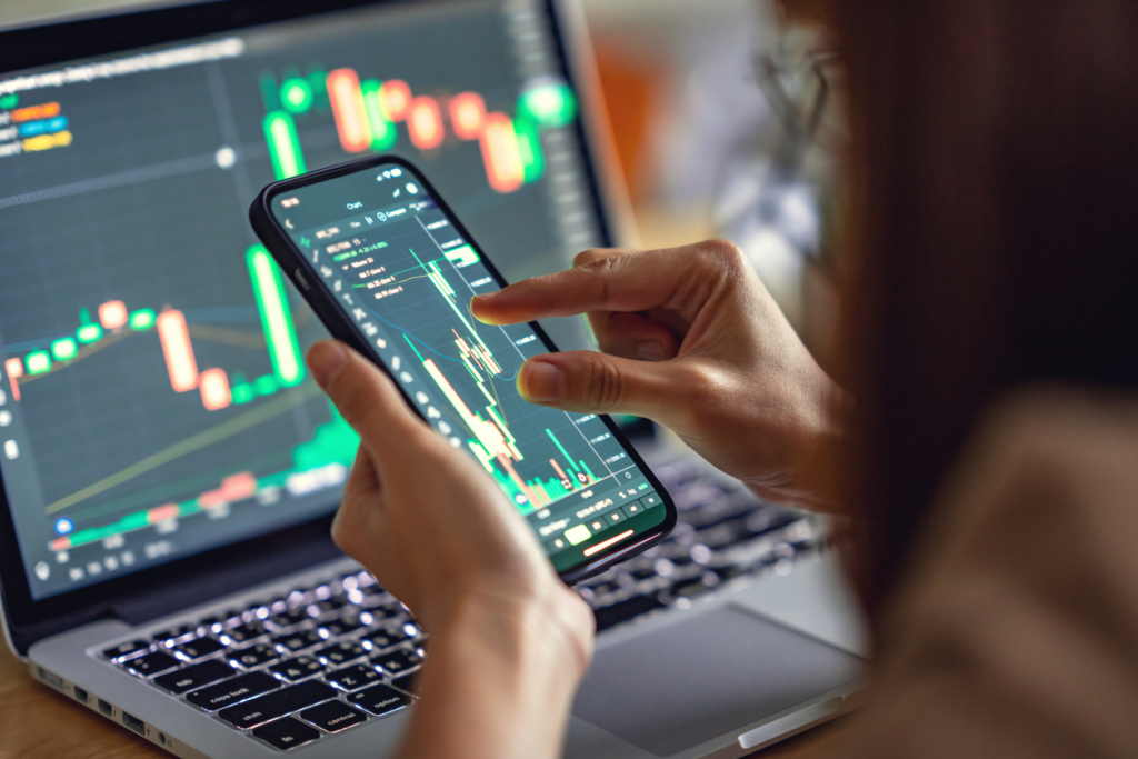Pessoa analisando gráficos de negociação financeira em um smartphone, com gráficos de alta e baixa no computador portátil ao fundo, focada em ações e mercado financeiro.