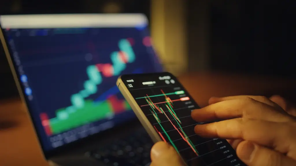 Pessoa analisando gráficos de ações no smartphone com fundo de computador com tela de análise de mercado financeiro, representando investimento em bolsa de valores.