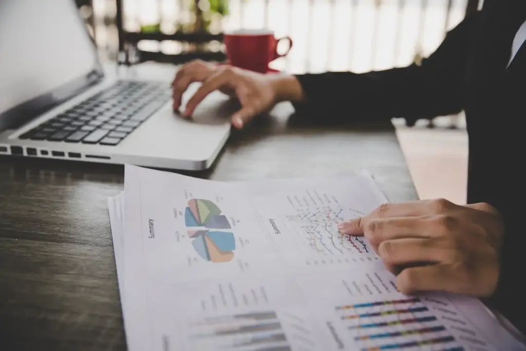 Pessoa analisando documentos financeiros e gráficos em uma mesa de trabalho, com laptop ao lado, no ambiente de escritório, promovendo produtividade.