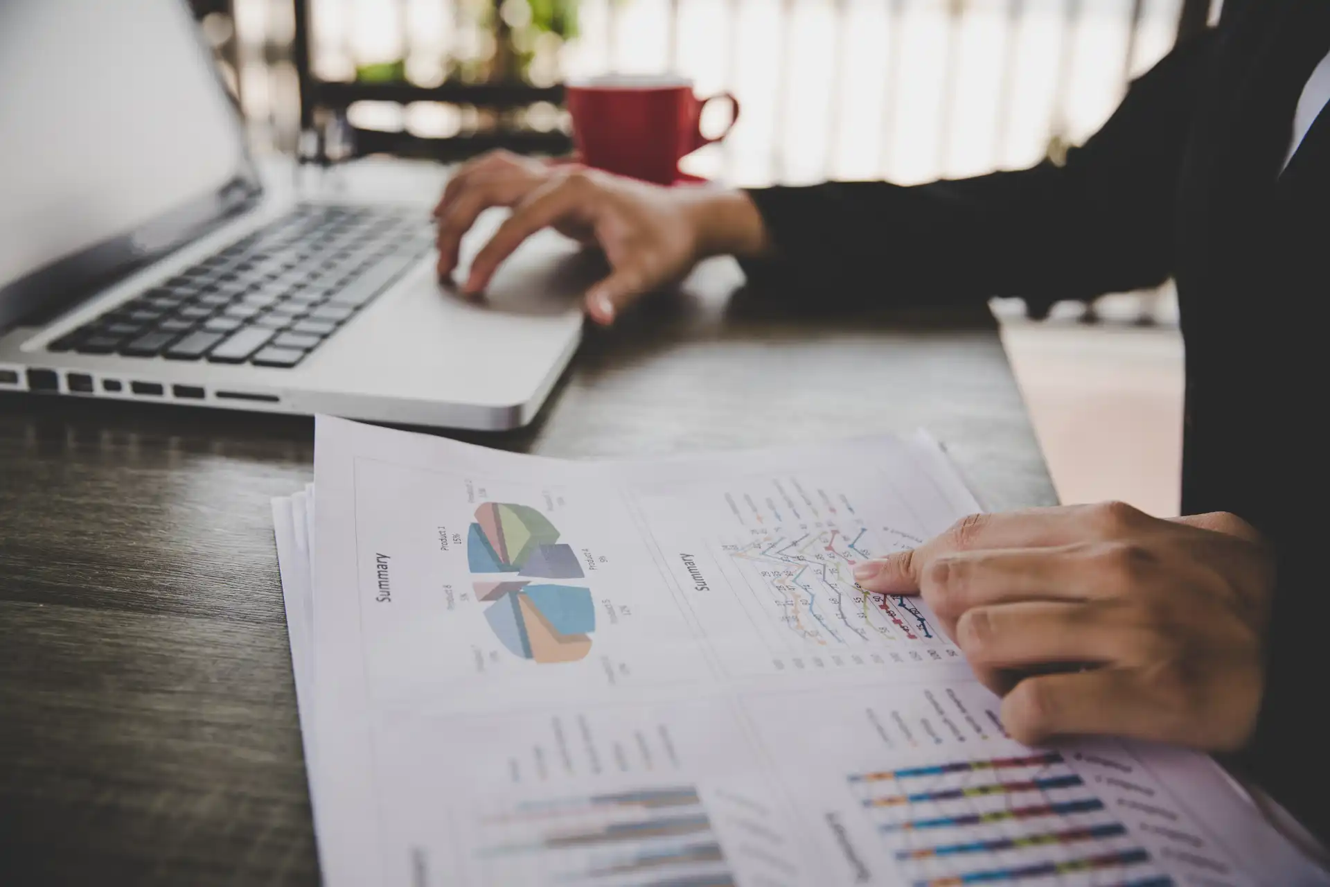 Pessoa analisando documentos financeiros e gráficos em uma mesa de trabalho, com laptop ao lado, no ambiente de escritório, promovendo produtividade.