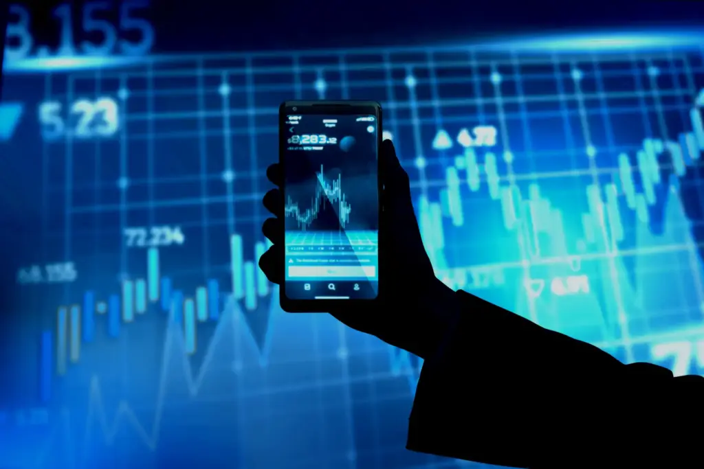 Pessoa segurando smartphone com gráfico de ações na tela, fundo com gráficos financeiros e dados do mercado financeiro, indicando análise de investimentos e trading.
