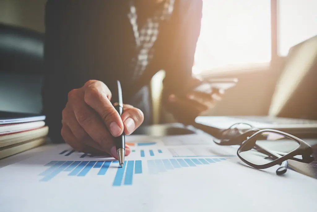 Pessoa analisando gráficos e dados financeiros em uma mesa de trabalho, com computador, óculos e documentos, em ambiente de escritório bem iluminado.