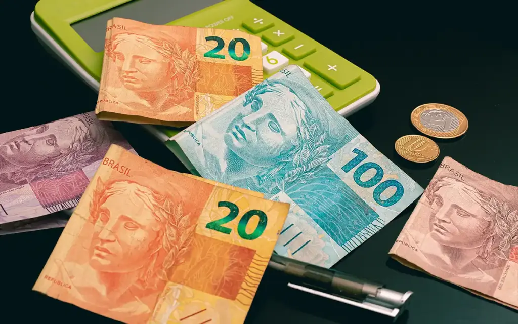 Notas de dinheiro do Brasil com cédulas de 20, 50 e 100 reais sobre um teclado de calculadora verde, com moedas e caneta ao lado, sugerindo cálculo e finanças pessoais.