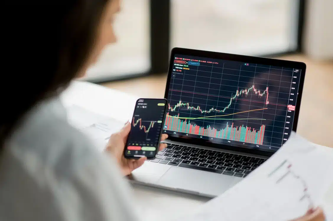 Pessoa analisando gráficos de ações de investimento em laptop e smartphone, focada em tendências financeiras e mercado acionário.