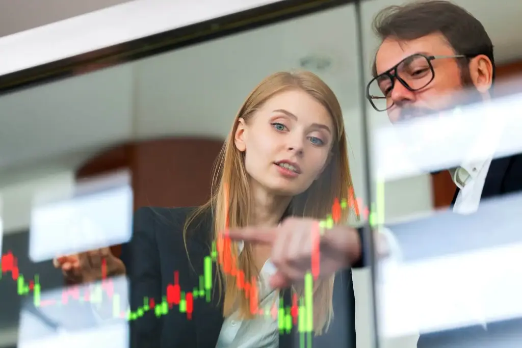 Homem e mulher analisando gráficos de ações na bolsa de valores, discutindo investimentos financeiros em um ambiente de negócios moderno.