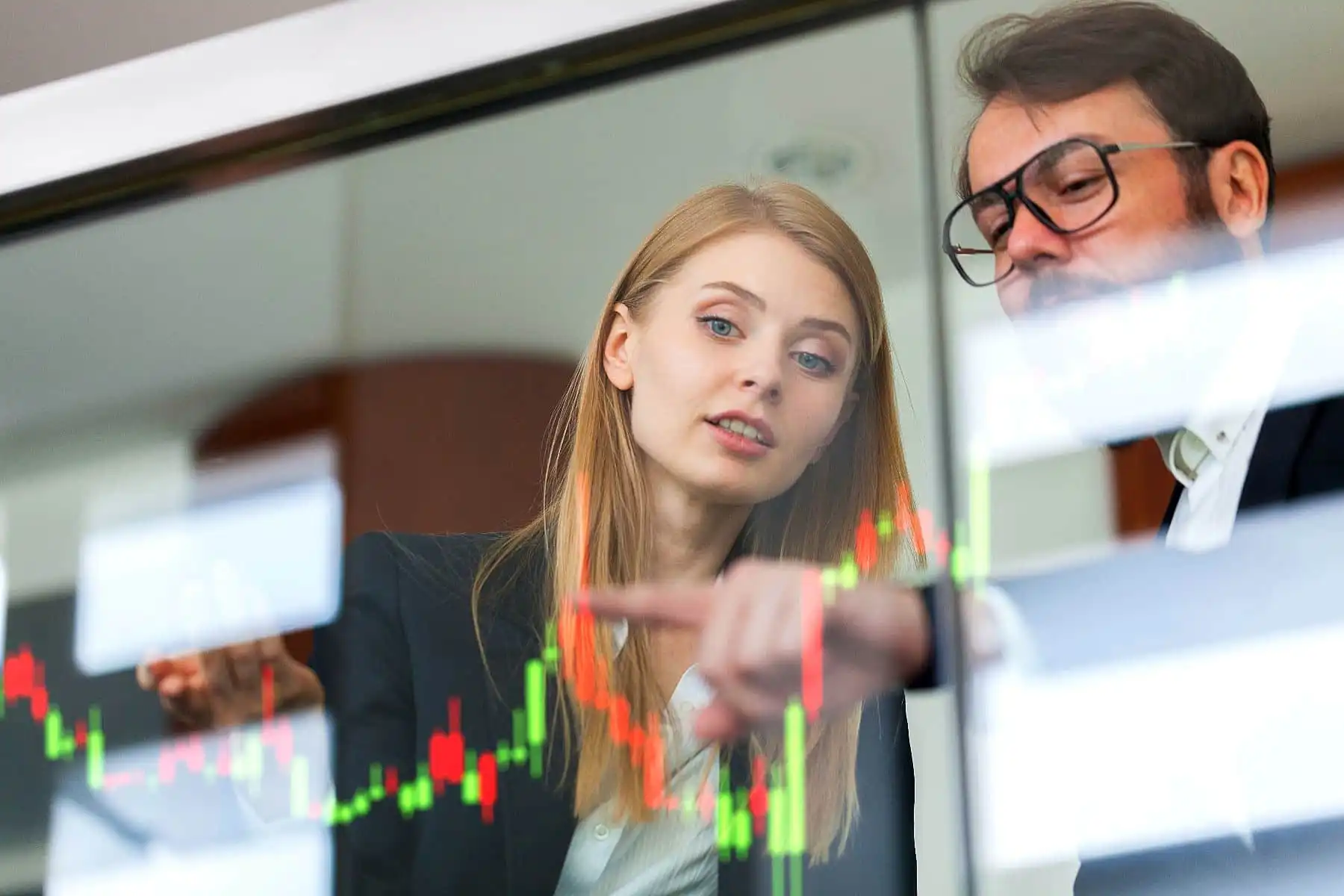 Homem e mulher analisando gráficos de ações na bolsa de valores, discutindo investimentos financeiros em um ambiente de negócios moderno.
