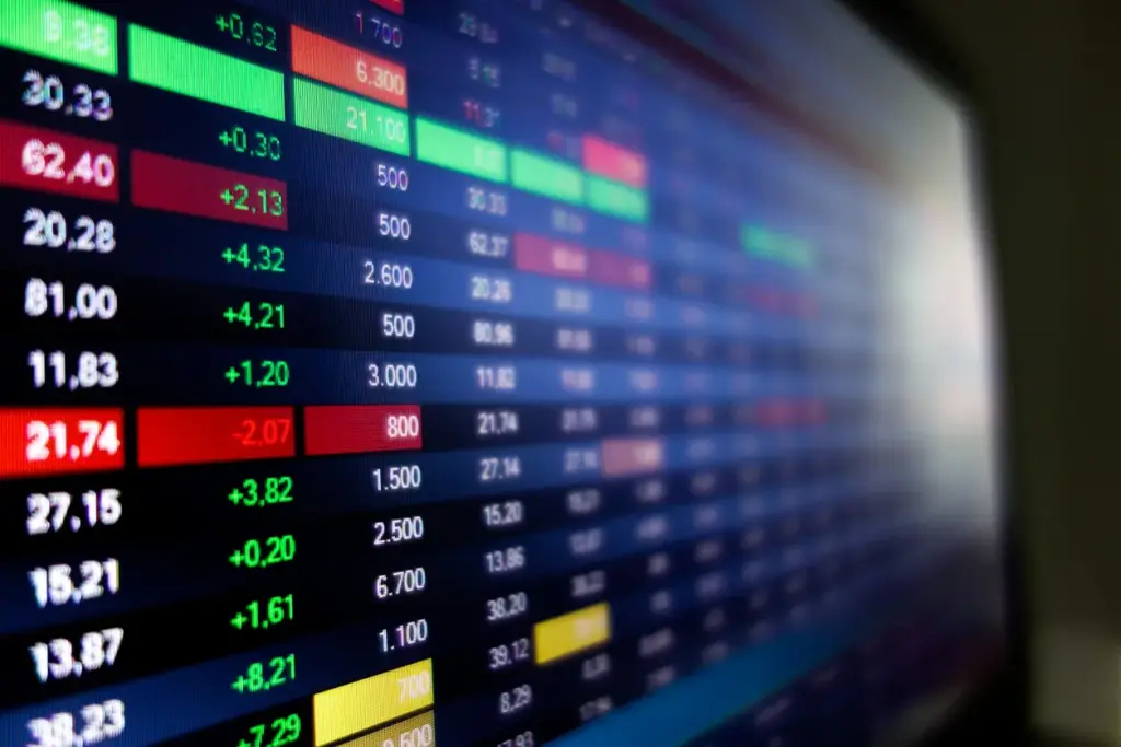 Tela de monitor em ambiente financeiro com gráficos e indicadores de preços, valores em vermelho e verde e números de variação em tempo real, focando no painel de cotações.